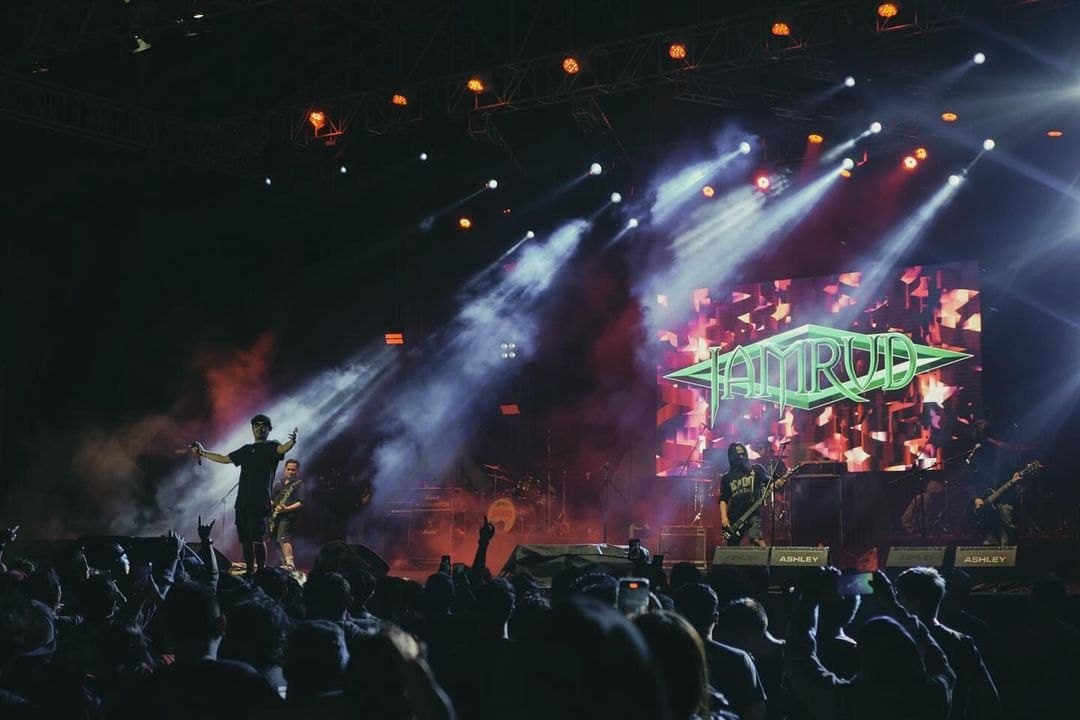 A concert stage with blue light beams and the band's name on the screen, seen over the crowd