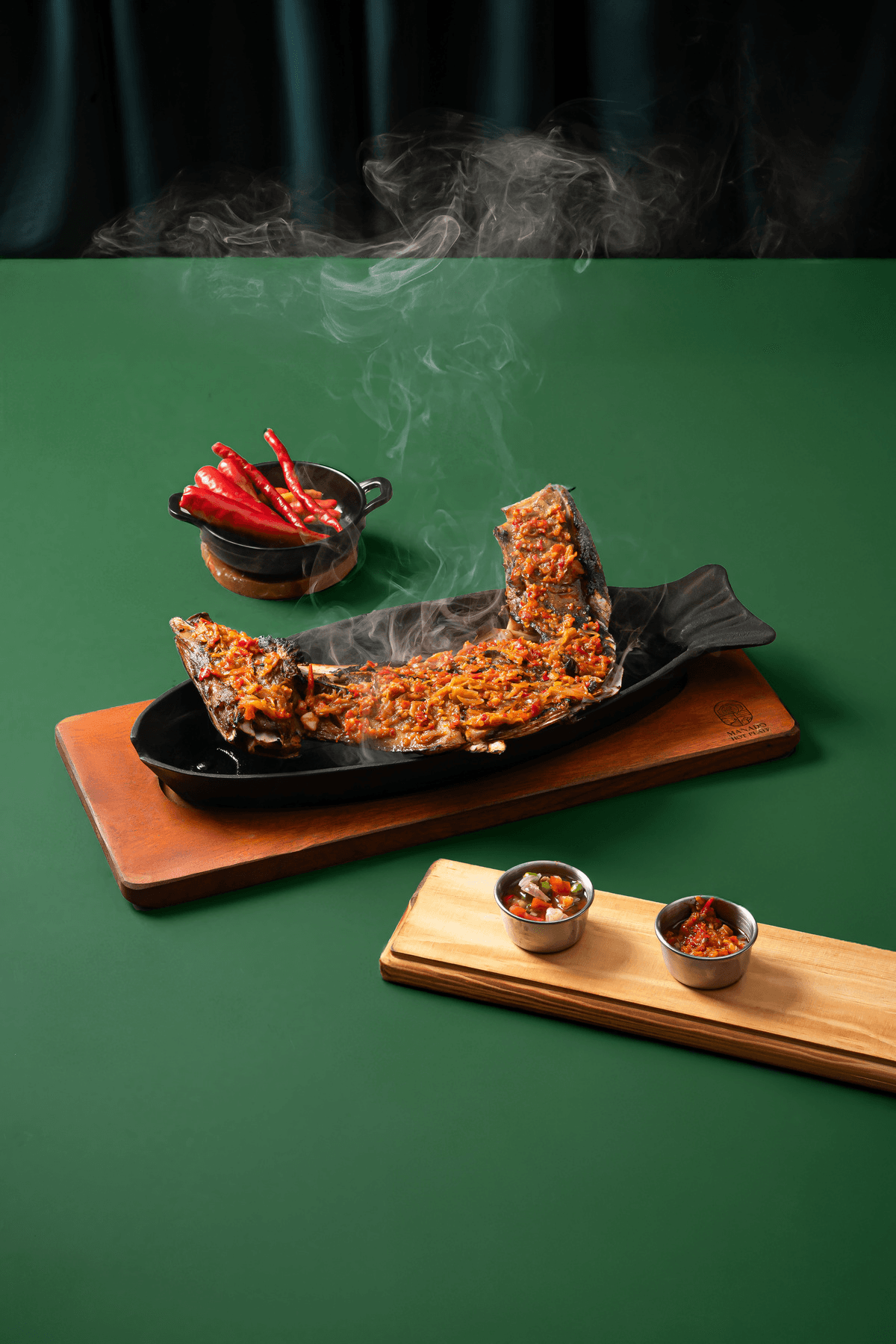 Grilled fish on a sizzling hot plate on a wooden board, chillies and sambal beside it, on a deep green background
