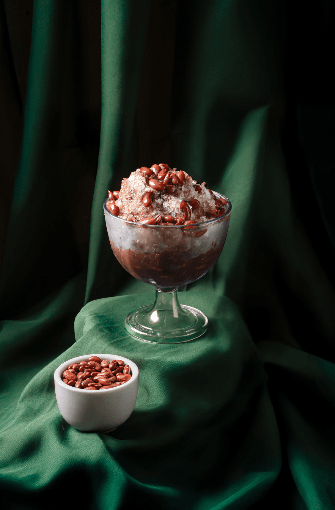 Shaved ice with red beans in a glass on folded green cloth, a small bowl of beans in front
