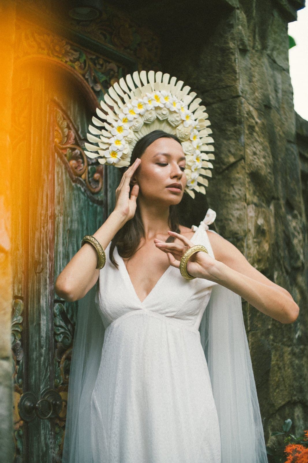 A woman in a white dress and a frangipani headdress in front of a carved stone wall