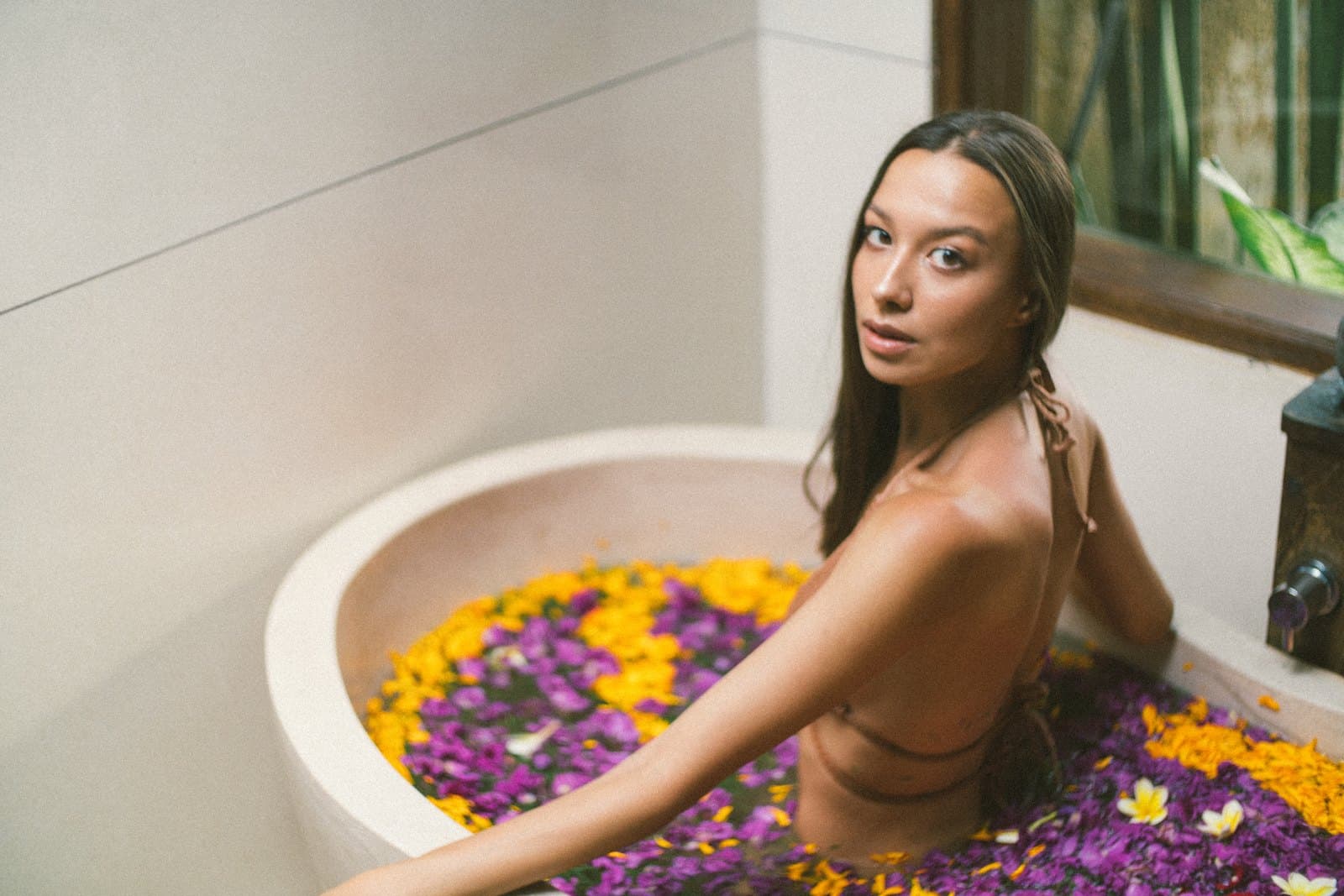 A woman in a stone bath filled with yellow and purple petals, green leaves behind