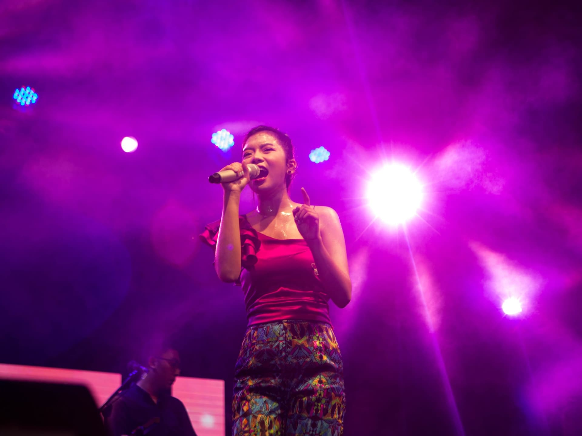 A singer in a red one-shoulder top and patterned trousers on stage under magenta light