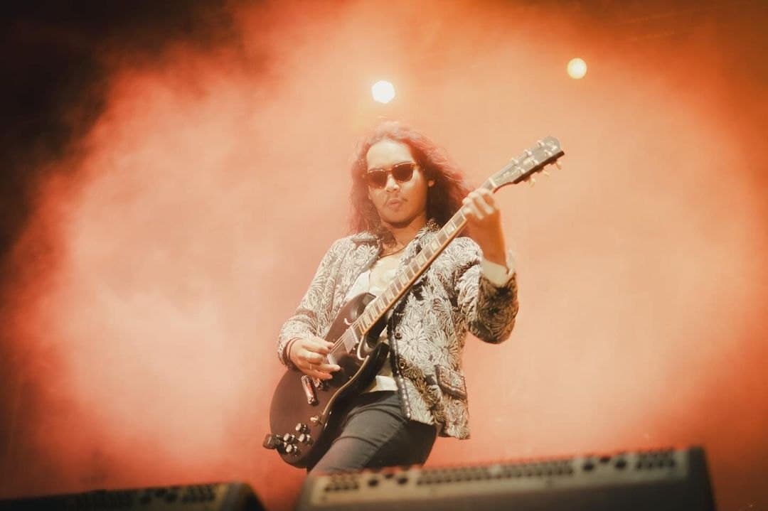 A long-haired guitarist in sunglasses and a patterned jacket in orange stage haze
