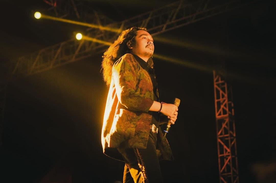 A long-haired vocalist in a patterned jacket singing under yellow light beside a stage truss