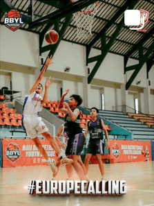 Feed tile: a player in white and blue laying up over two defenders under the gym roof, Bali Basketball Youth League banners on the court