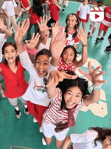 Feed tile, reel: children in red and white cheering with their arms up, seen from above on a green court