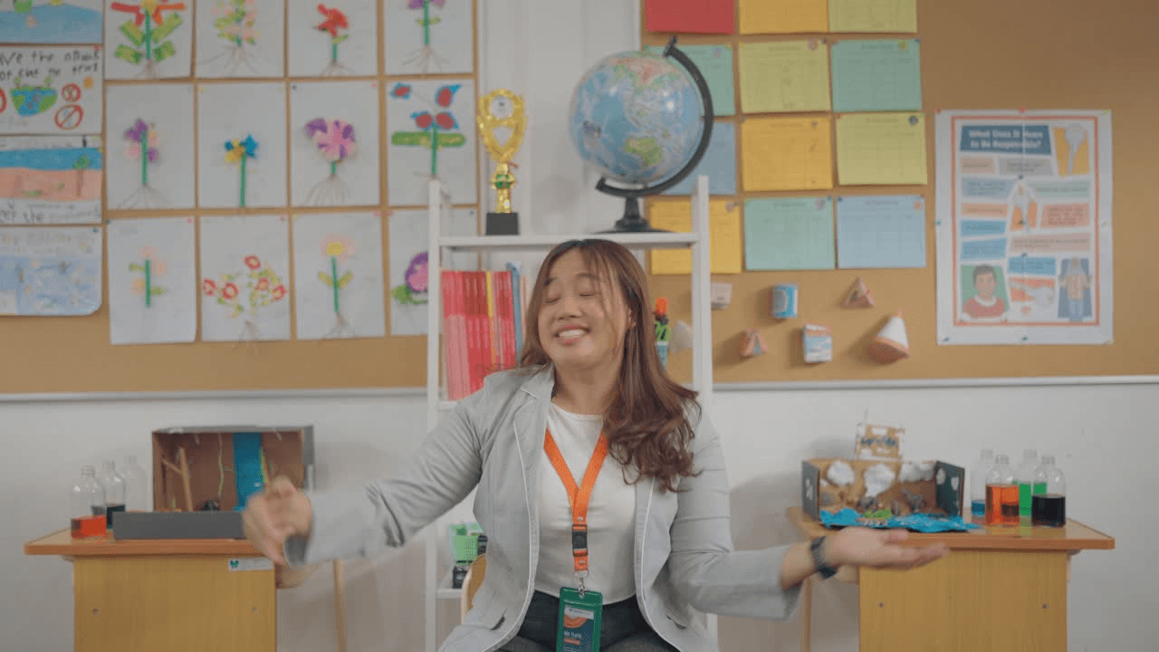 A teacher at a desk in a classroom with a globe and children's drawings